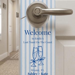 May include: A rectangular door hanger with blue and white vertical stripes. The text "Welcome to Emily's Last Toast On The Coast" is in navy blue. Below are two champagne flutes and the names Ashley + Kate.