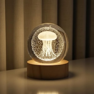 Peut inclure: Une sculpture de méduse lumineuse enfermée dans une sphère de verre transparente, éclairée par des lumières internes. La méduse est blanche, avec des tentacules délicats. La sphère repose sur une base en bois clair et ronde, créant une lumière ambiante chaleureuse.