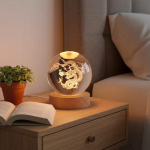 Può includere: Lampada a sfera di cristallo con un design a drago dorato. La lampada è posizionata su una base di legno ed emette una luce calda. Si trova su un comodino di legno, accanto a una pianta e a un libro aperto.