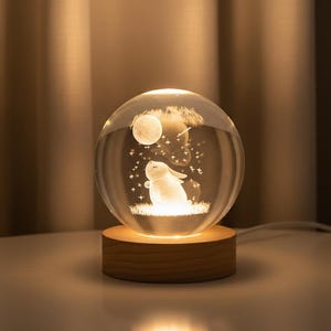 May include: A clear glass sphere featuring an illuminated 3D design of a rabbit, moon, and stars. The sphere rests on a wooden base, giving off a warm light. The product is a decorative night light.