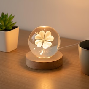 Può includere: Una luce notturna a sfera di vetro luminoso con un design a quadrifoglio. La sfera poggia su una base rotonda in legno. La luce emette un bagliore soffuso, illuminando il quadrifoglio e le stelle. È visibile una piccola pianta succulenta in un vaso bianco.