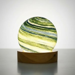 Puede incluir: Un globo decorativo de vidrio con patrones arremolinados de verde, azul y amarillo, apoyado sobre una base redonda de madera. El globo está iluminado desde dentro, creando un brillo suave. El diseño es moderno y artístico.