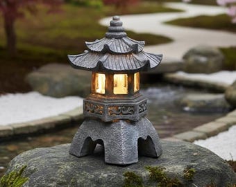 May include: A gray stone pagoda-style garden lantern with a multi-tiered roof and decorative carvings. The lantern has a warm, inviting glow from the interior light. It sits on a large, moss-covered rock in a serene garden setting.