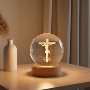 May include: A clear glass sphere featuring a detailed depiction of Jesus on the cross, illuminated from within. The sphere rests on a round, wooden base. A white vase with dried flowers is to the left.