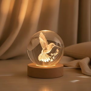 May include: A clear glass sphere with a 3D hummingbird design, illuminated from within. The sphere sits on a round, light-colored wooden base. The hummingbird is depicted with outstretched wings and a long beak, perched on a branch.