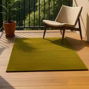 May include: A rectangular, olive green rug sits on a wooden deck, with a beige chair and potted plant in the background. The rug's texture is visible, and the scene is bathed in sunlight, suggesting an outdoor setting.