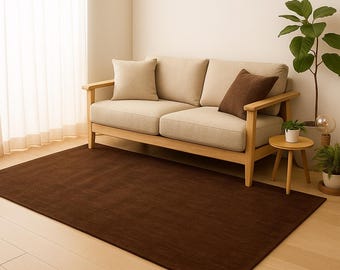 May include: A rectangular, dark brown rug is placed on a light wood floor in front of a beige sofa with wooden arms. The sofa has two beige cushions and a brown pillow. A small wooden side table and potted plants are to the right.