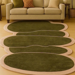 May include: A modern olive green area rug with a unique, organic shape. The rug features a light beige border and a thin white accent line. The rug is placed on a light wood floor, with a beige sofa and pillows in the background.