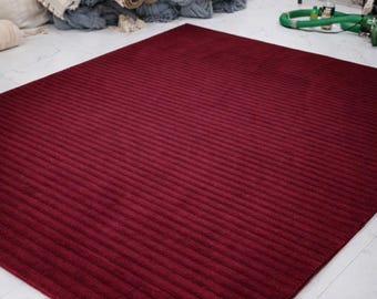 Puede incluir: Una alfombra grande, rectangular, de color burdeos con una textura acanalada. La alfombra está colocada sobre un suelo de baldosas blancas. En el fondo hay rollos de tela y un montón de material azul.