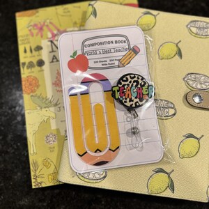 May include: A teacher-themed gift set featuring a pencil-shaped paper clip, a badge reel with the word "TEACHER", and a composition notebook. The notebook has the text "World's Best Teacher" and a red apple illustration.