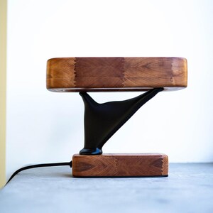 Puede incluir: Una lámpara de madera moderna con una parte superior e inferior rectangulares, conectadas por un soporte negro estilizado. Los componentes de madera de la lámpara muestran una veta de madera cálida y natural. Un cable de alimentación negro sale de la base.