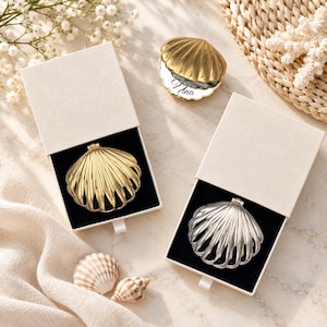Könnte beinhalten: Zwei muschelförmige Kompaktspiegel, einer in Gold und einer in Silber, jeweils in einer weißen Schiebeschachtel mit schwarzem Innenfutter. Ein dritter goldener Muschelspiegel mit dem eingravierten Namen Nina liegt oberhalb der Schachteln.