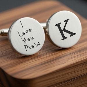 May include: A pair of silver cufflinks with a round face. One cufflink has the text "I Love You More" in a script font. The other cufflink has a black letter "K". The cufflinks are displayed on a wooden surface.