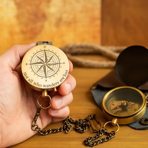 May include: A vintage brass compass with a chain is held in a hand. The compass has a detailed compass rose design and the inscription "Nor all who wanders are lost". A second compass with a black face and open lid is also visible.