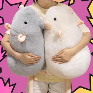 May include: Two large, plush rat-shaped cushions. One is light grey, the other is white. Both have small pink ears, noses, and tongues. The cushions are held against a pink and yellow background.