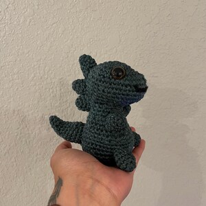 May include: A handmade, dark teal crochet dinosaur with large, brown eyes. The amigurumi-style creature has a spiky back and a small, smiling mouth. The toy is held in a hand, showcasing the detailed craftsmanship of the plushie.