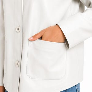 May include: A cream-coloured jacket with a classic design. The jacket has a button-up front, two front pockets, and a tailored fit. The jacket is made of a smooth material. The model is wearing blue jeans.