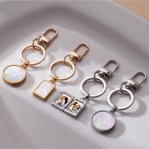 Op de afbeelding: Vier sleutelhangers met verschillende ontwerpen. Twee hebben goudkleurige hardware, een met een ronde hanger en de andere met een rechthoekige hanger. De andere twee sleutelhangers hebben zilverkleurige hardware, een met een ronde hanger en de andere met een kleine fotolijst.