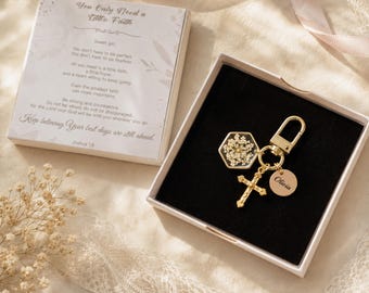 May include: A gold-toned keychain featuring a hexagonal resin charm with white pressed flowers, a cross pendant, and a small round disc engraved with the name Olivia. The keychain is presented in a square box with a card printed with religious text.