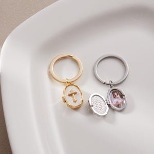 May include: Two keyrings with oval pendants. One is gold-toned with a white enamel cross pendant. The other is silver-toned with an oval locket pendant, open to show a photo. Both have round key rings.