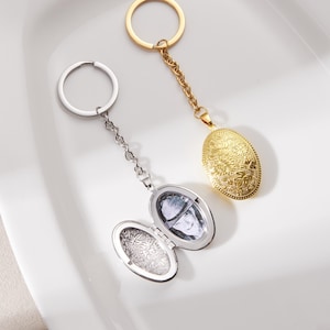 Op de afbeelding: Twee ovale medaillon sleutelhangers. Eén is zilverkleurig, open om een foto te onthullen, en de andere is goudkleurig. Beide hebben decoratieve bloempatronen en zijn bevestigd aan sleutelringen. De gouden sleutelhanger is gesloten.