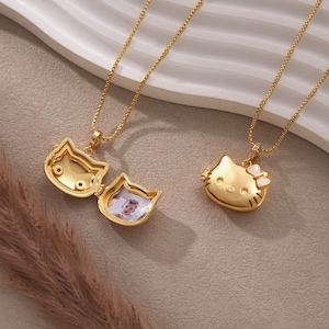 May include: Two gold-colored cat-shaped locket necklaces. One locket is open, revealing a small photo inside. The other locket features a white bow. Both necklaces have delicate gold chains, perfect for a gift.