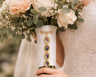 Può includere: Un bouquet da sposa con rose color crema e pesca, nebbiolina e fogliame di eucalipto. Un nastro di raso bianco avvolge i gambi, decorato con un ciondolo verticale in metallo dorato con quattro piccole cornici ovali per fotografie.