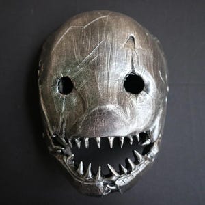 May include: A silver-colored mask with a distressed finish, featuring large eye holes and a wide, toothy grin. The mask has a textured surface with visible cracks and scratches, giving it a weathered appearance. The mask is likely for costume or decorative purposes.