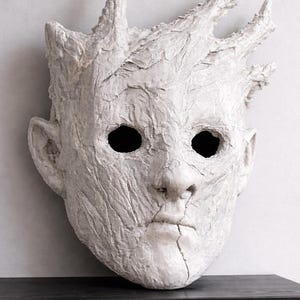 Puede incluir: Una escultura de máscara blanca texturizada con rostro humano y protuberancias en forma de corona. La máscara tiene cuencas oculares oscuras y una superficie agrietada, montada sobre una estantería negra contra una pared neutra.
