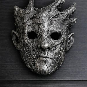 Puede incluir: Una máscara plateada con una apariencia texturizada y desgastada. La máscara presenta un rostro humanoide con cuencas oculares vacías y una superficie detallada y agrietada, que se asemeja a la corteza de un árbol. La parte superior de la máscara tiene un diseño similar a una corona.