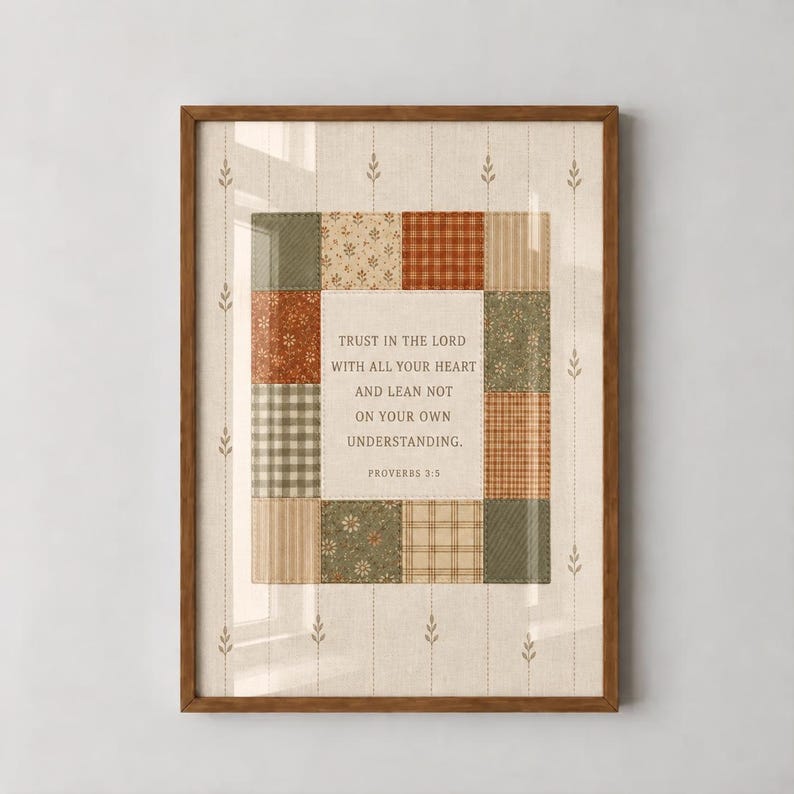 Op de afbeelding: Een ingelijste wandkunstprint met een centraal wit paneel met de tekst Trust in the Lord with all your heart and lean not on your own understanding. Proverbs 3:5, omringd door een patchworkrand van groene, beige en oranje stofpatronen.
