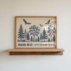 May include: A framed wall art print featuring a forest landscape with dark blue pine trees and three eagles in flight against a cream background. The text reads: ISAIAH 40:31 | THEY WILL SOAR ON WINGS LIKE EAGLES. The print is displayed on a wooden shelf.