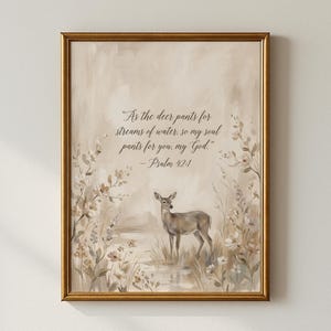 May include: A framed wall art print featuring a deer standing in a field of flowers with a stream. The text reads: "As the deer pants for streams of water, so my soul pants for you, my God." - Psalm 42:1. The artwork uses neutral beige tones.