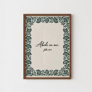 May include: A rectangular wall art print in a brown wooden frame featuring a cream-coloured background with a dark green floral border. The centre text reads "Abide in me. John 15:4" in a black script and serif font, centred within the floral frame.