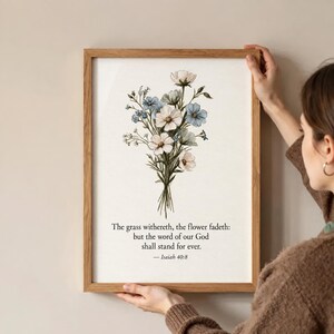 May include: A framed wall art print featuring a watercolour illustration of a bouquet of white and blue flowers. Below the floral arrangement, the text reads: The grass withereth, the flower fadeth: but the word of our God shall stand for ever. — Isaiah 40:8.