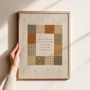 May include: A framed wall art print featuring a central square block of patchwork fabric patterns in shades of olive green, rust orange, and cream. The center contains the text: Trust in the Lord with all your heart and lean not on your own understanding. Proverbs 3:5.