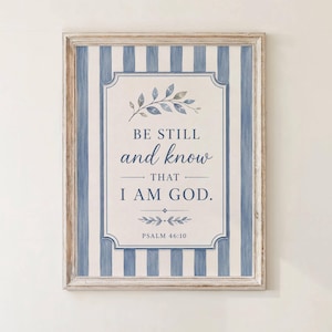 May include: A framed wall art print featuring a blue and white vertical striped background. The center contains a decorative border with the text "BE STILL and know THAT I AM GOD." and "PSALM 46:10" in blue, accented by small leaf illustrations.