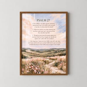 May include: A framed wall art print featuring Psalm 27 text overlaid on a landscape painting of a dirt path winding through a field of pink wildflowers and rolling hills under a soft, pastel-coloured sky with clouds in shades of peach and blue.