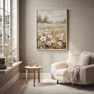 May include: A framed painting depicts a figure walking in a field of flowers. The artwork features a soft color palette of pink, white, and green, with a light brown wooden frame. The painting hangs above a cream-colored armchair.