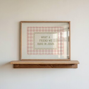May include: A framed wall art piece featuring a light pink and white gingham pattern background with a central rectangular panel containing the text "WHAT A FRIEND WE HAVE IN JESUS" in a simple, dark-colored, sans-serif typeface.
