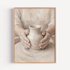 May include: A framed print shows hands moulding a light-coloured ceramic vase on a pottery wheel. The artwork is rendered in soft, neutral tones, with a focus on the hands and the vase. The print is set in a light-coloured wooden frame.
