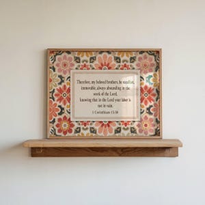 May include: A rectangular wooden frame containing a print with a floral pattern in shades of orange, yellow, and green. The centre features a white rectangular panel with the text: Therefore, my beloved brothers, be steadfast, 1 Corinthians 15:58.