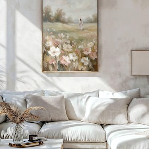 May include: A framed floral landscape painting with a person in white walking through a field of flowers. The painting has a wooden frame and is displayed above a white sectional sofa with beige pillows. A coffee table with a vase and candle sits in front of the sofa.