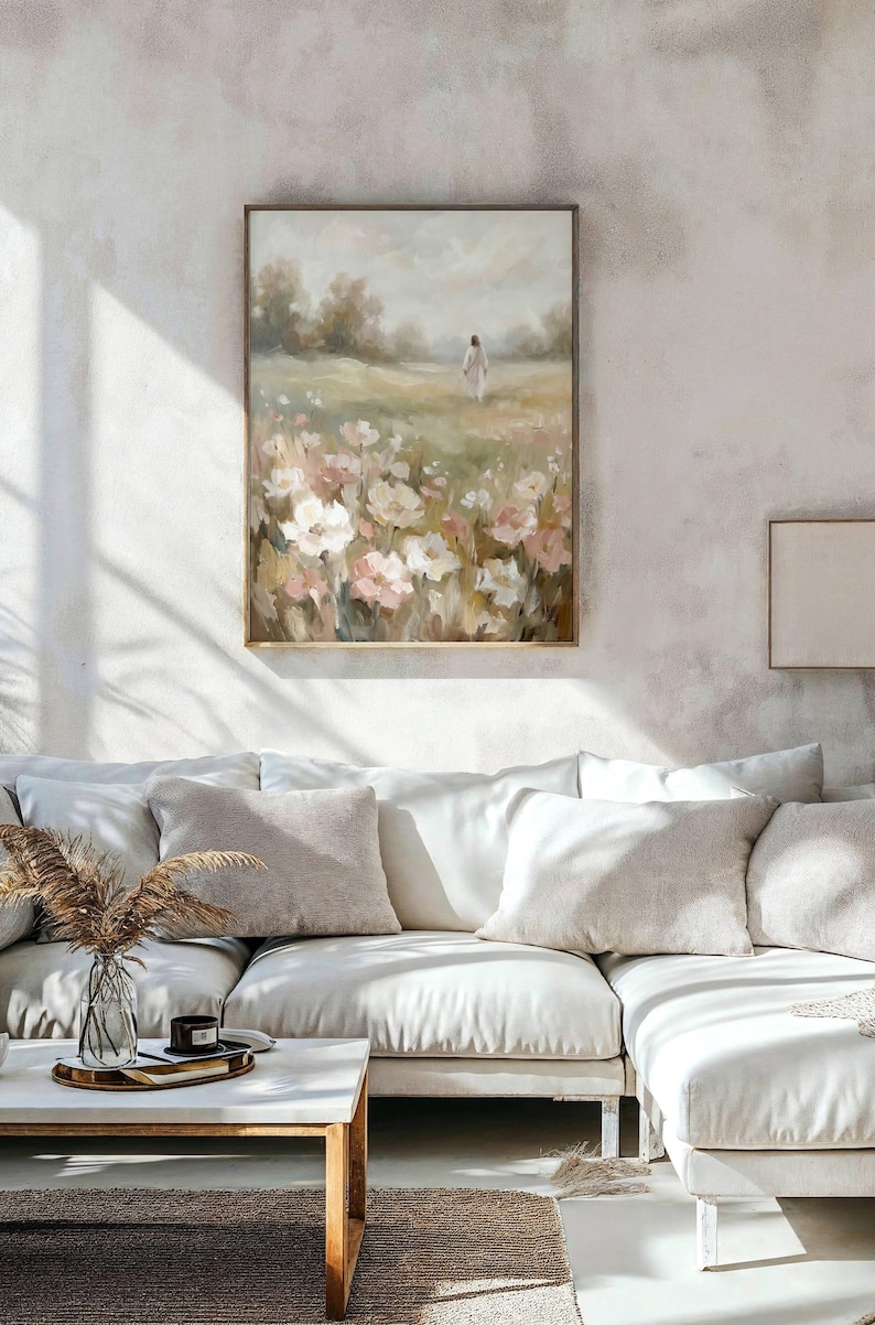 May include: A framed floral landscape painting with a person in white walking through a field of flowers. The painting has a wooden frame and is displayed above a white sectional sofa with beige pillows. A coffee table with a vase and candle sits in front of the sofa.