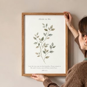 Könnte beinhalten: Ein rechteckiger Holzrahmen mit einem weißen Druck, der eine Aquarellillustration eines grünen Blattzweigs zeigt. Der Text lautet oben Abide in Me und unten I am the vine; you are the branches. If you remain in Me and I in you, you will bear much fruit. John 15:5.