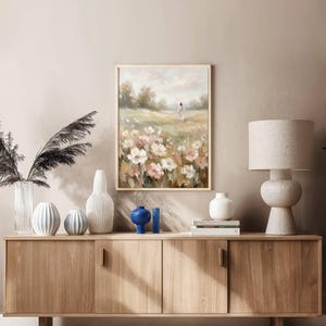 May include: A framed floral landscape painting with a person walking in a field of white and pink flowers. The painting hangs above a wooden cabinet with various decorative vases and a lamp.