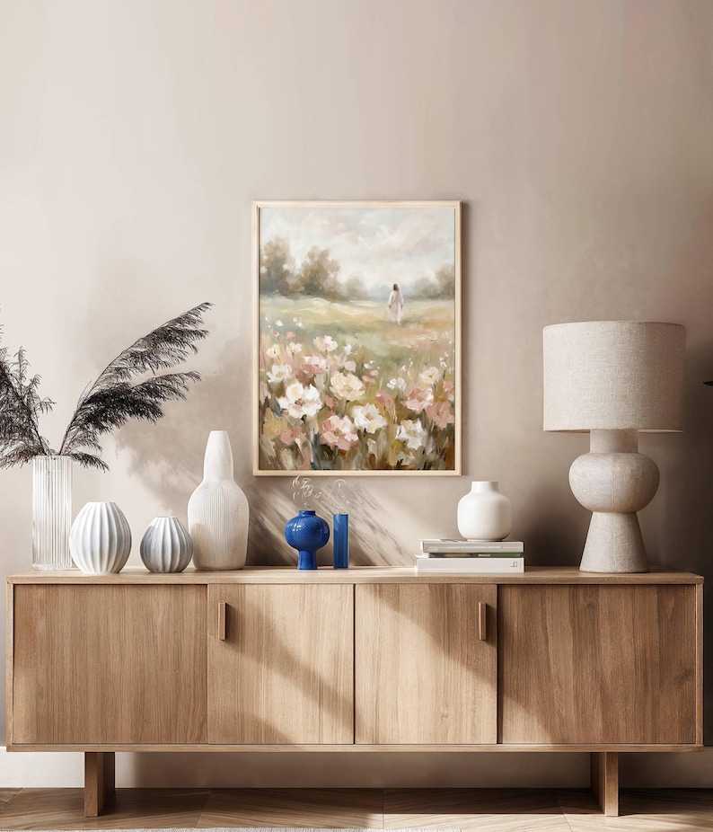 May include: A framed floral landscape painting with a person walking in a field of white and pink flowers. The painting hangs above a wooden cabinet with various decorative vases and a lamp.