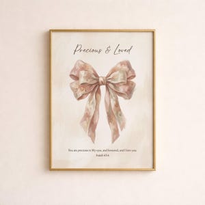 Pode incluir: Uma impressão emoldurada com uma ilustração em aquarela de um grande laço rosa com detalhes florais. O texto "Precious & Loved" está acima do laço, com o versículo bíblico Isaías 43:4 abaixo. A moldura é dourada.