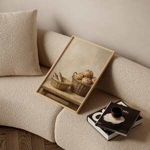 May include: A framed print depicting two woven baskets, one with fish and the other with round loaves of bread. The artwork is set against a neutral background and rests on a light-colored sofa, with books and a cup nearby.
