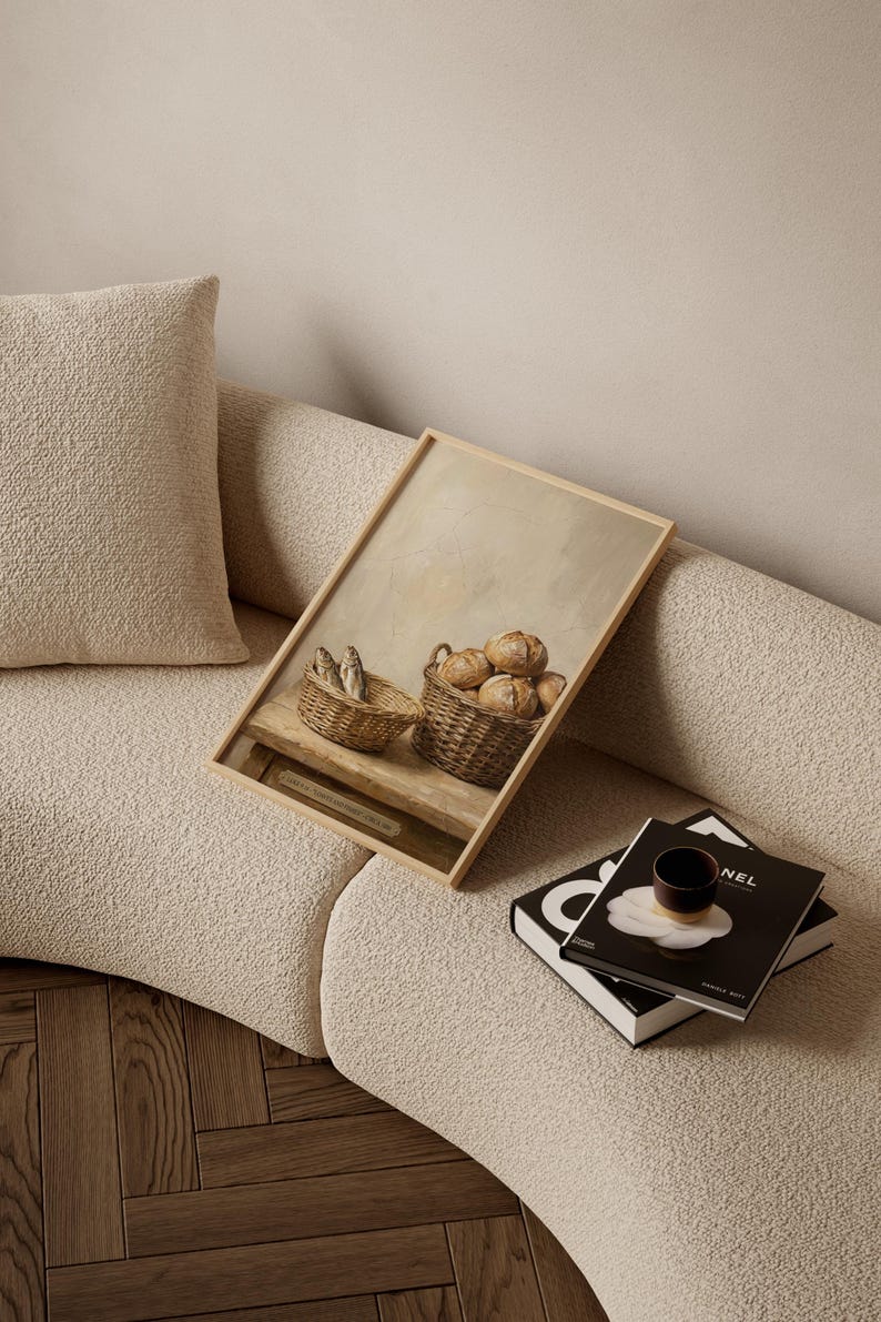 May include: A framed print depicting two woven baskets, one with fish and the other with round loaves of bread. The artwork is set against a neutral background and rests on a light-colored sofa, with books and a cup nearby.